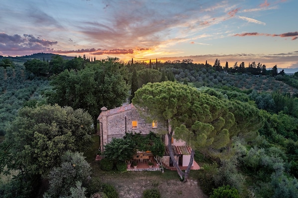A rustic stone house surrounded by dense greenery, with a clear view of the sunset in the background.