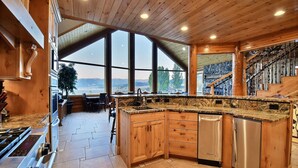 A spacious kitchen with large windows, a stone backsplash, wooden cabinets, and a stainless steel dishwasher.