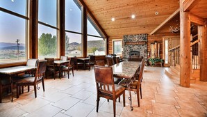 A spacious dining area with a large table and chairs, a stone fireplace, and large windows offering a view of the outdoors.