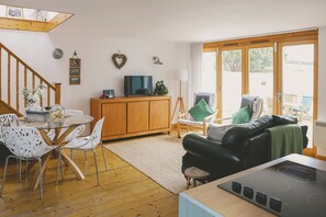 A living room with a wooden staircase, a dining area with white chairs, a black leather sofa, and a kitchen with a stove. - Delightful retreat, 3 mins walk to Widemouth Bay blue flag beaches & coast path (Widemouth Bay)