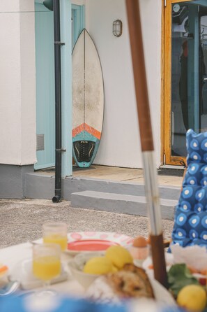 A surfboard leaning against a wall, a window with a view, and a table with food items. - Delightful retreat, 3 mins walk to Widemouth Bay blue flag beaches & coast path (Widemouth Bay)