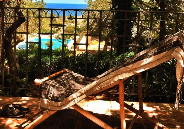 Una cadira de sol amb coixí rayat, situada en una zona amb ombra i vista a una piscina i vegetació.