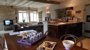 A spacious kitchen with a dining area, featuring a wooden table set with pastries and glasses, a television in the background, and a stone wall. - Beautiful stone house "country chic", private heated pool, play area (Membrey)