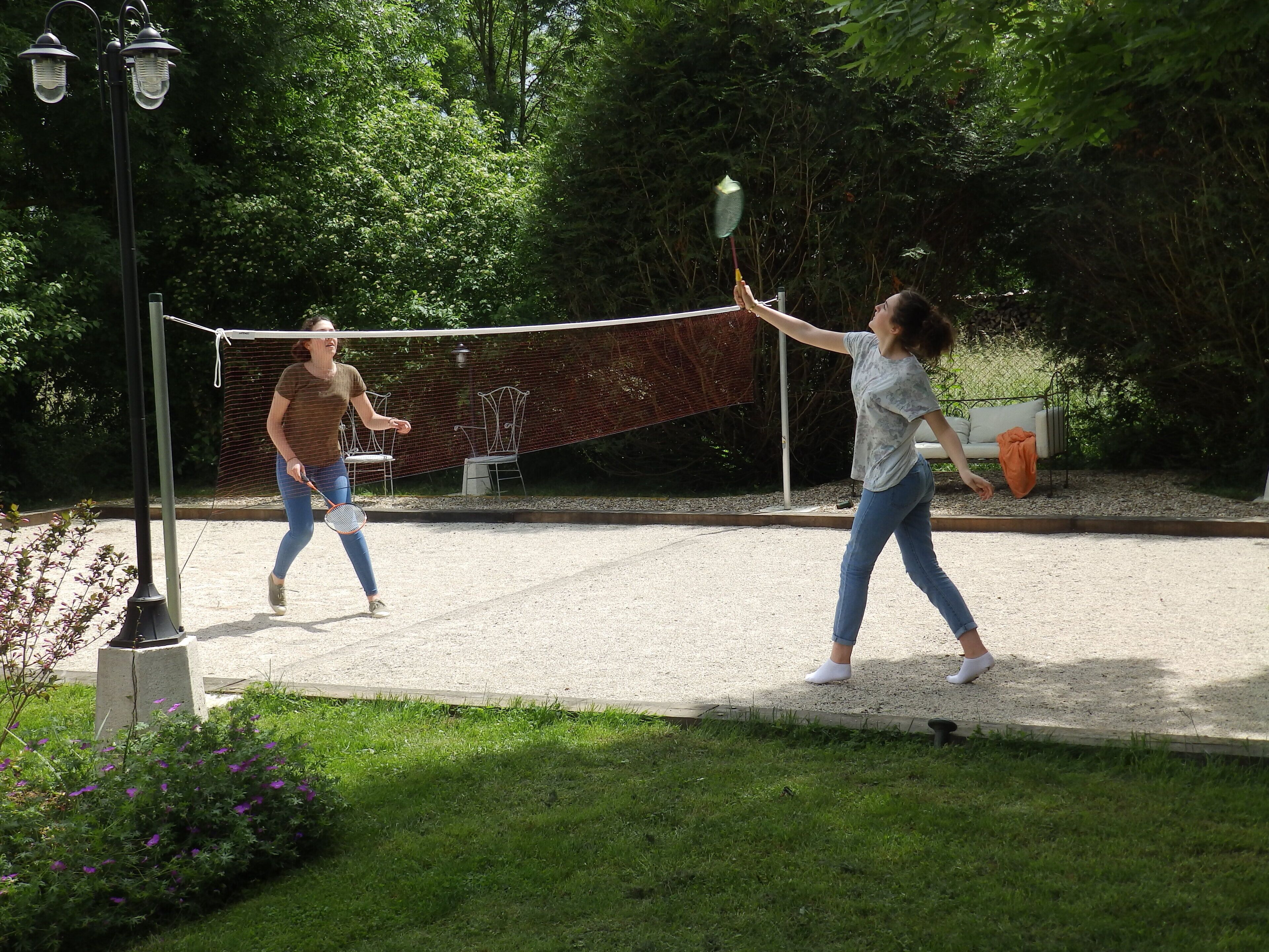 Two people playing badminton on a gravel court with a fence and trees in the background.