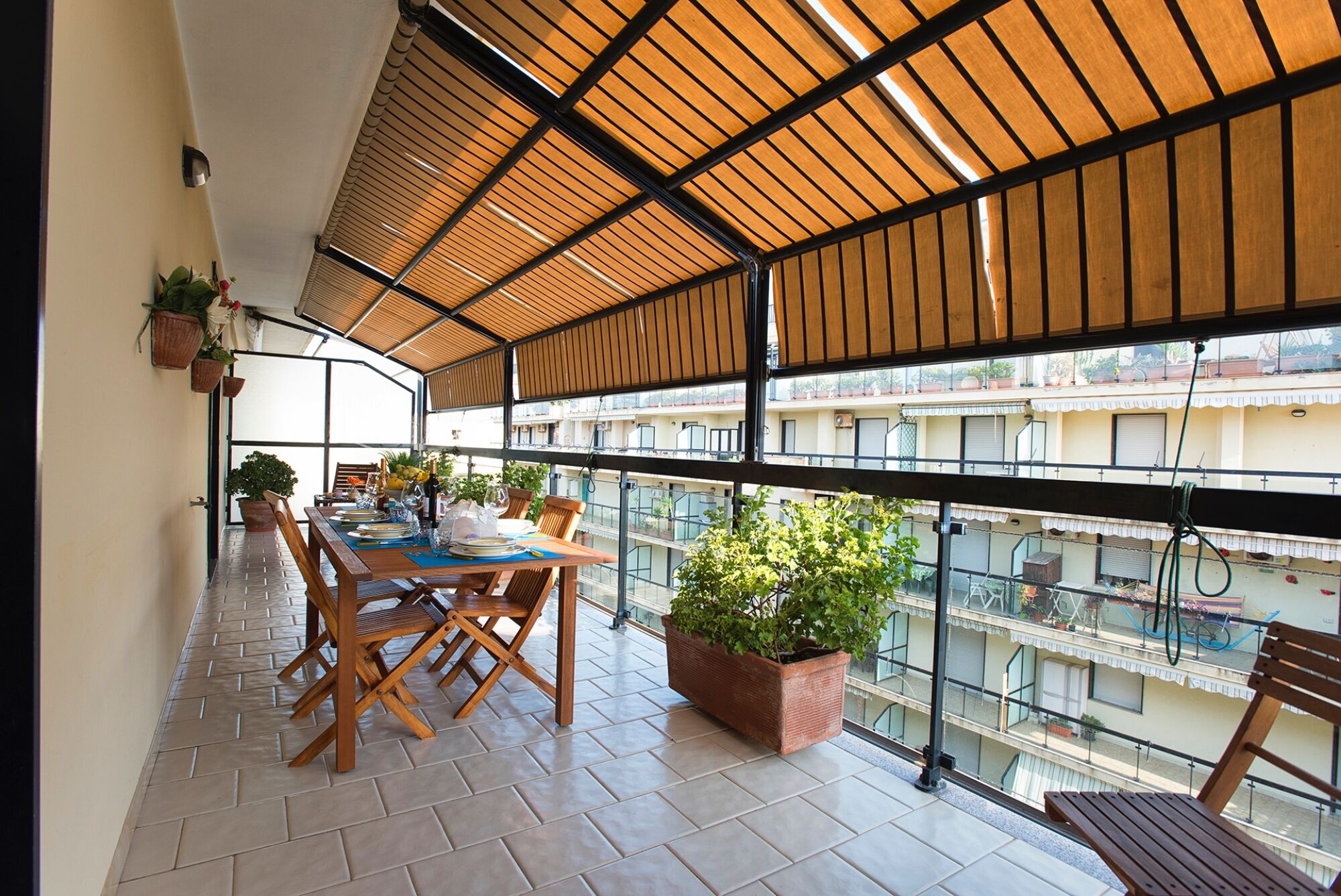 Uma varanda com mesa de madeira posta para refeições, plantas em vasos e vista para a paisagem urbana.