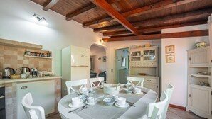 A dining area with a round table set for four, surrounded by white chairs. A kitchen area with a refrigerator and cabinets is visible in the background. - Villa Kala Nostra by the sea, 150m and 5m walk from Kal'eMoru/Villasimius shoreline (Geremeas)