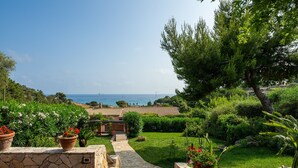 A garden with a stone pathway leading to a house with a tiled roof, surrounded by lush greenery and a view of the sea. - Villa Kala Nostra by the sea, 150m and 5m walk from Kal'eMoru/Villasimius shoreline (Geremeas)