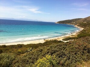 A coastal landscape with a beach, clear blue waters, and a hilly backdrop.