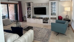 Un salon avec un canapé blanc, un fauteuil bleu, un téléviseur à écran plat, une bibliothèque et une petite table avec une lampe.