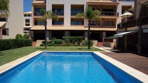 Une piscine rectangulaire bordée de carreaux, entourée d’une terrasse en bois. Au fond, on aperçoit un immeuble à plusieurs étages doté de balcons, ainsi qu’un jardin planté de palmiers et d’arbustes.