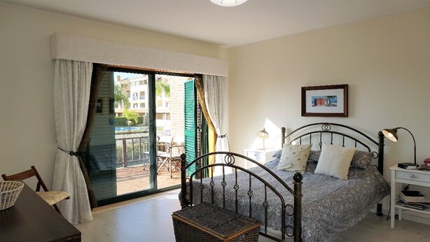 Une chambre à coucher comprenant un lit, une table de chevet, une lampe, une chaise, une fenêtre avec des rideaux et un balcon donnant sur des immeubles.