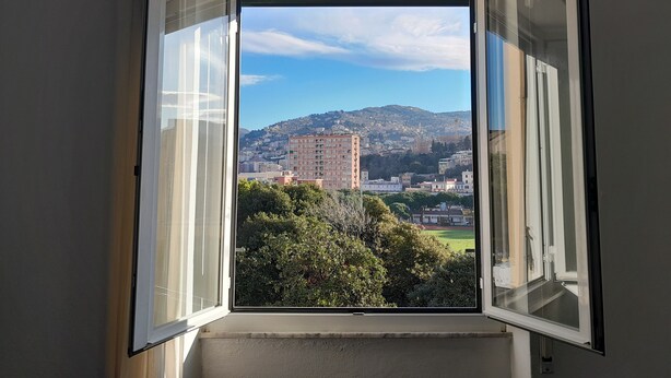 Una habitación con ventana abierta que muestra un paisaje urbano con edificios y una colina.