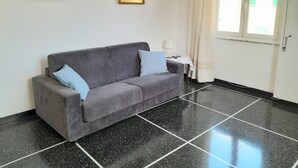 Una habitación con un sofá gris, dos almohadas, una mesita con una lámpara y dos cuadros enmarcados en la pared.
