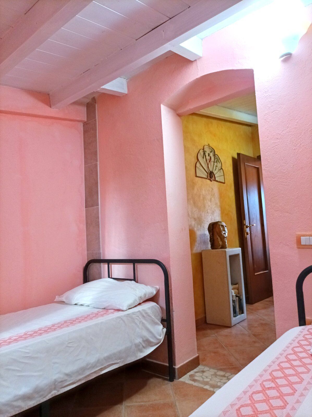A small, single-bed room with pink walls, a tiled floor, and a wooden door.