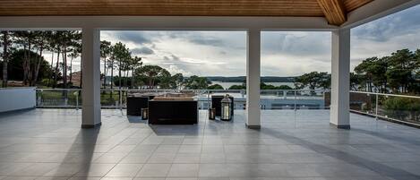 A spacious terrace with a view of a lake and trees, featuring a wooden ceiling and tiled flooring.