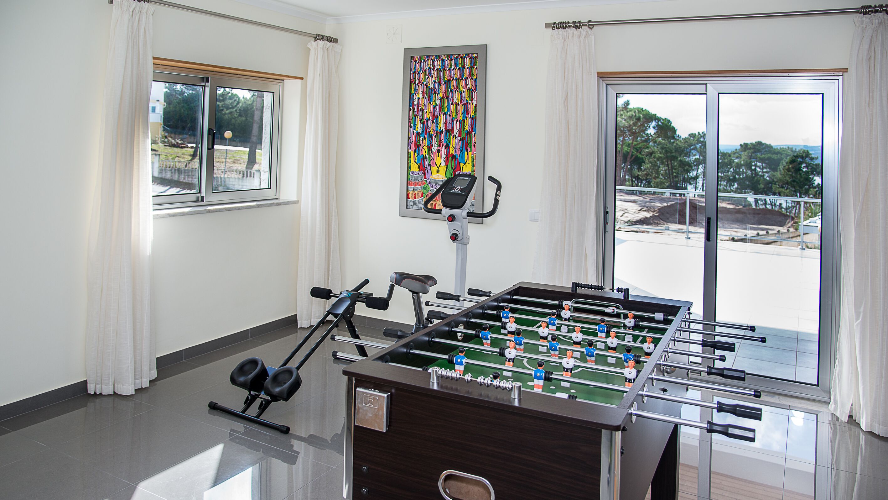 A room with a foosball table, exercise equipment, and large windows with curtains.
