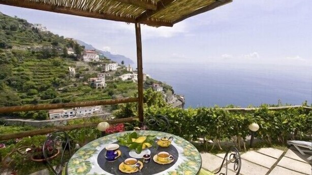 A round table with a floral tablecloth, surrounded by chairs, set on a terrace overlooking a coastal landscape with buildings and greenery. - Mariuccia House Amalfi (Amalfi)