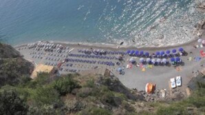 A beach with rows of blue umbrellas and sun loungers, a rocky shoreline, and a few people in the distance. - Mariuccia House Amalfi (Amalfi)