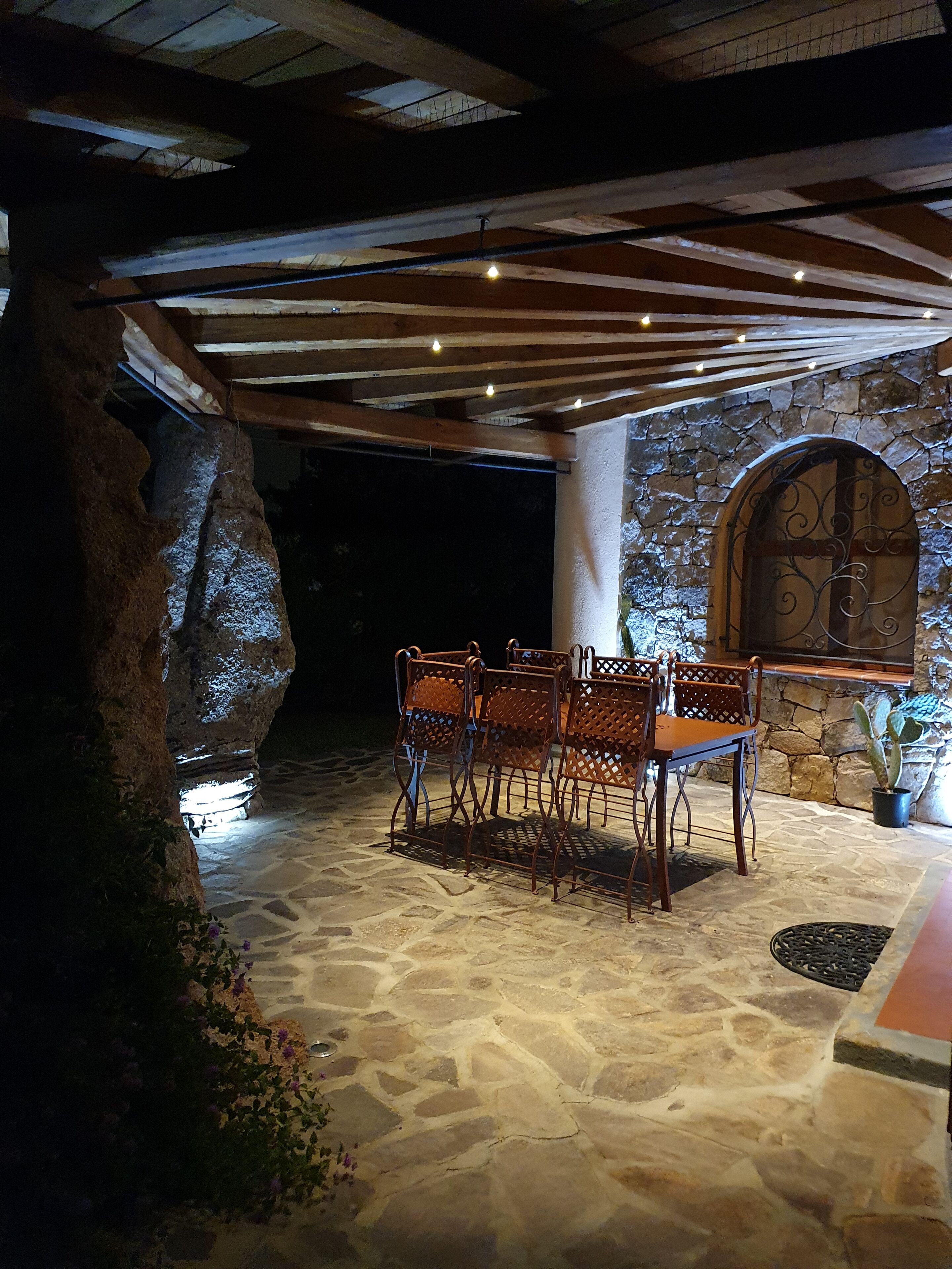 A stone patio with a table and chairs, a stone wall with an arched window, and a wooden ceiling structure.