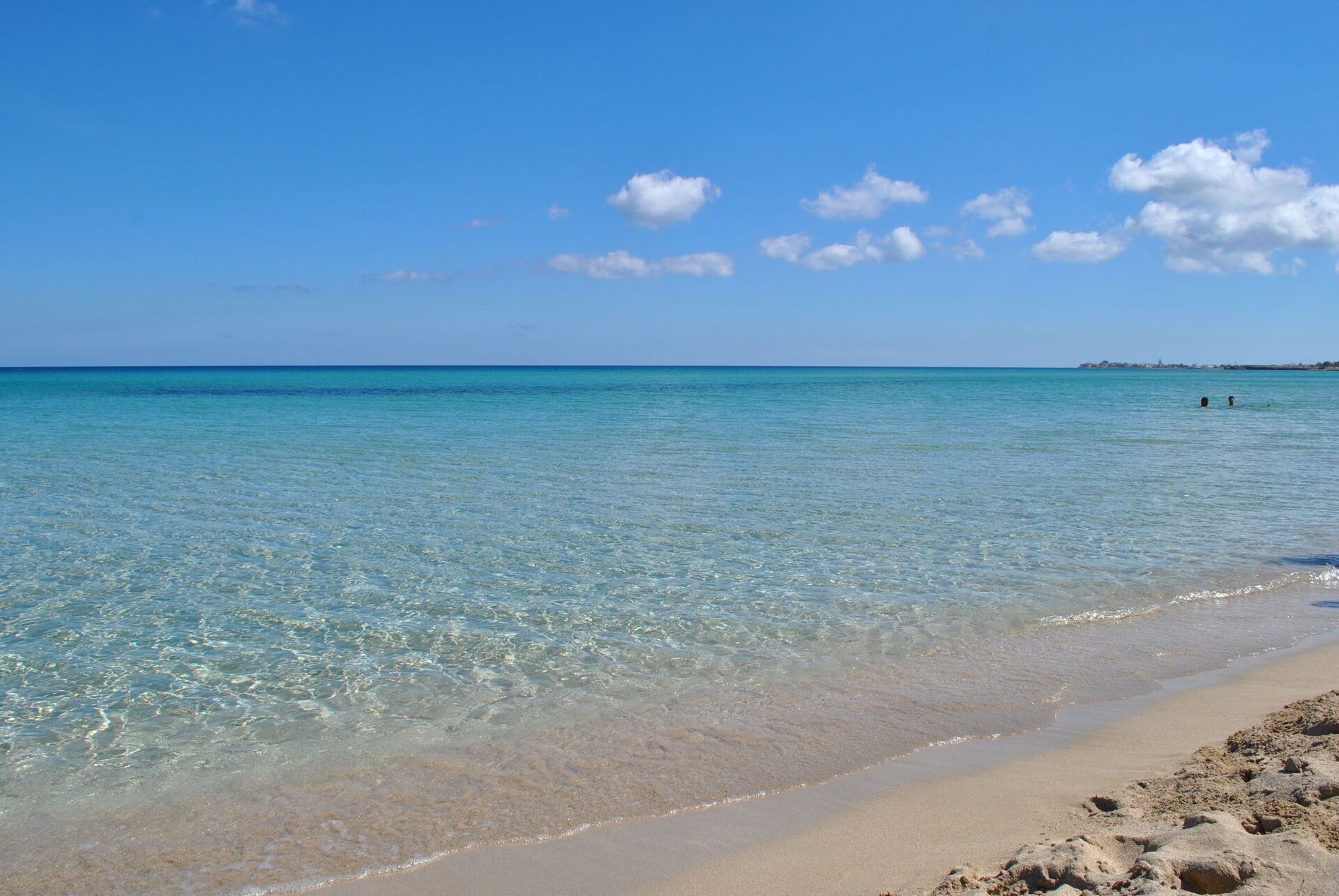 Una platja amb aigua turquesa cristal·lina, sorra daurada i una línia de costa llunyana amb edificis.