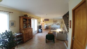 Ein geräumiges Wohnzimmer mit einem Holzschrank, einem Fernseher und einem grünen Sessel. Im Hintergrund ist ein Essbereich mit Tisch und Stühlen zu sehen.