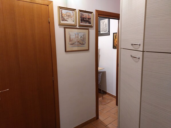 Ein schmaler Korridor mit Holztüren und weißen Wänden. An der Wand hängen drei gerahmte Bilder, und im Hintergrund ist ein weißer Kühlschrank zu sehen.