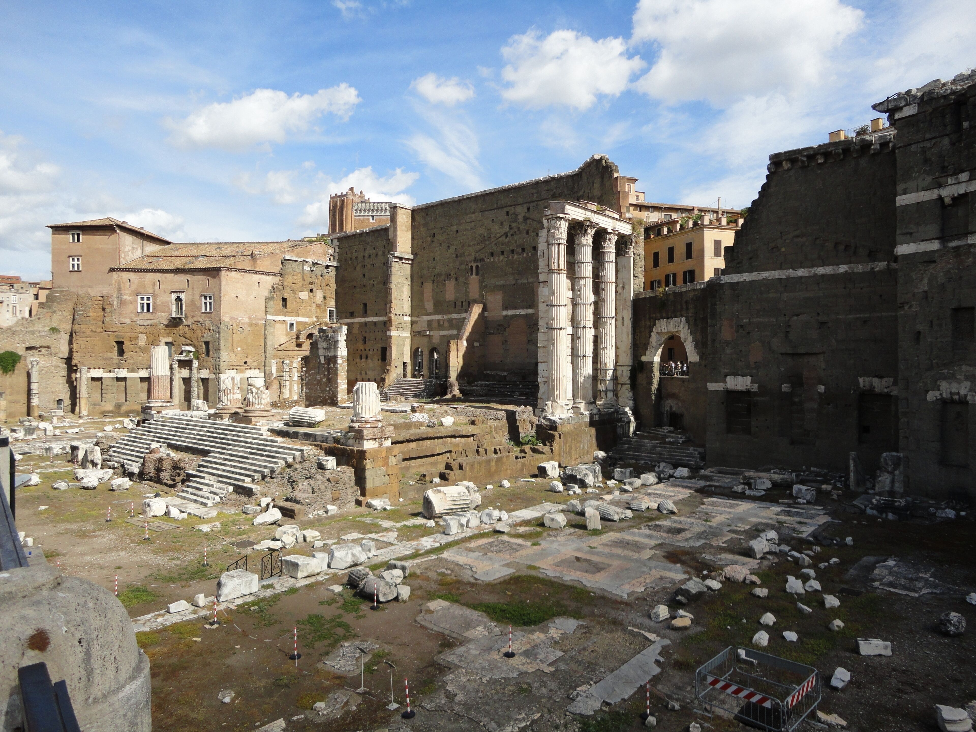 Ancient ruins with columns and steps, surrounded by modern buildings.