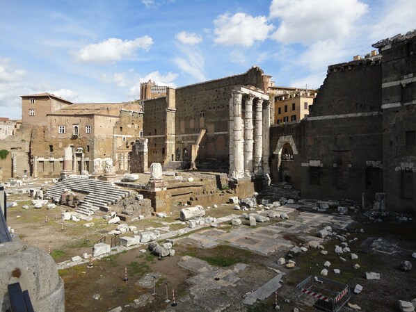 Ancient ruins with columns and steps, surrounded by modern buildings.