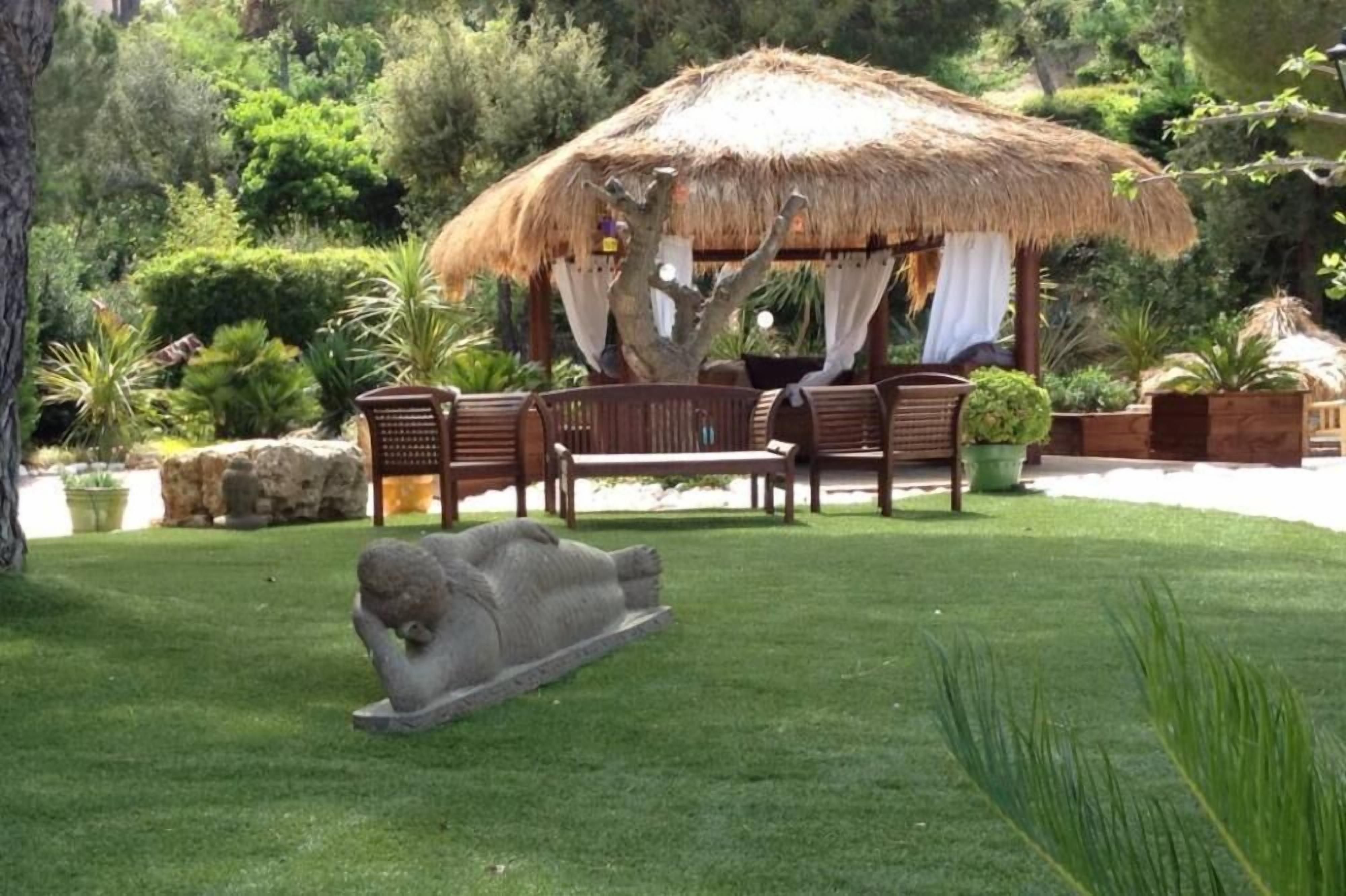 A thatched-roof gazebo with wooden furniture and a stone sculpture in a well-manicured garden.