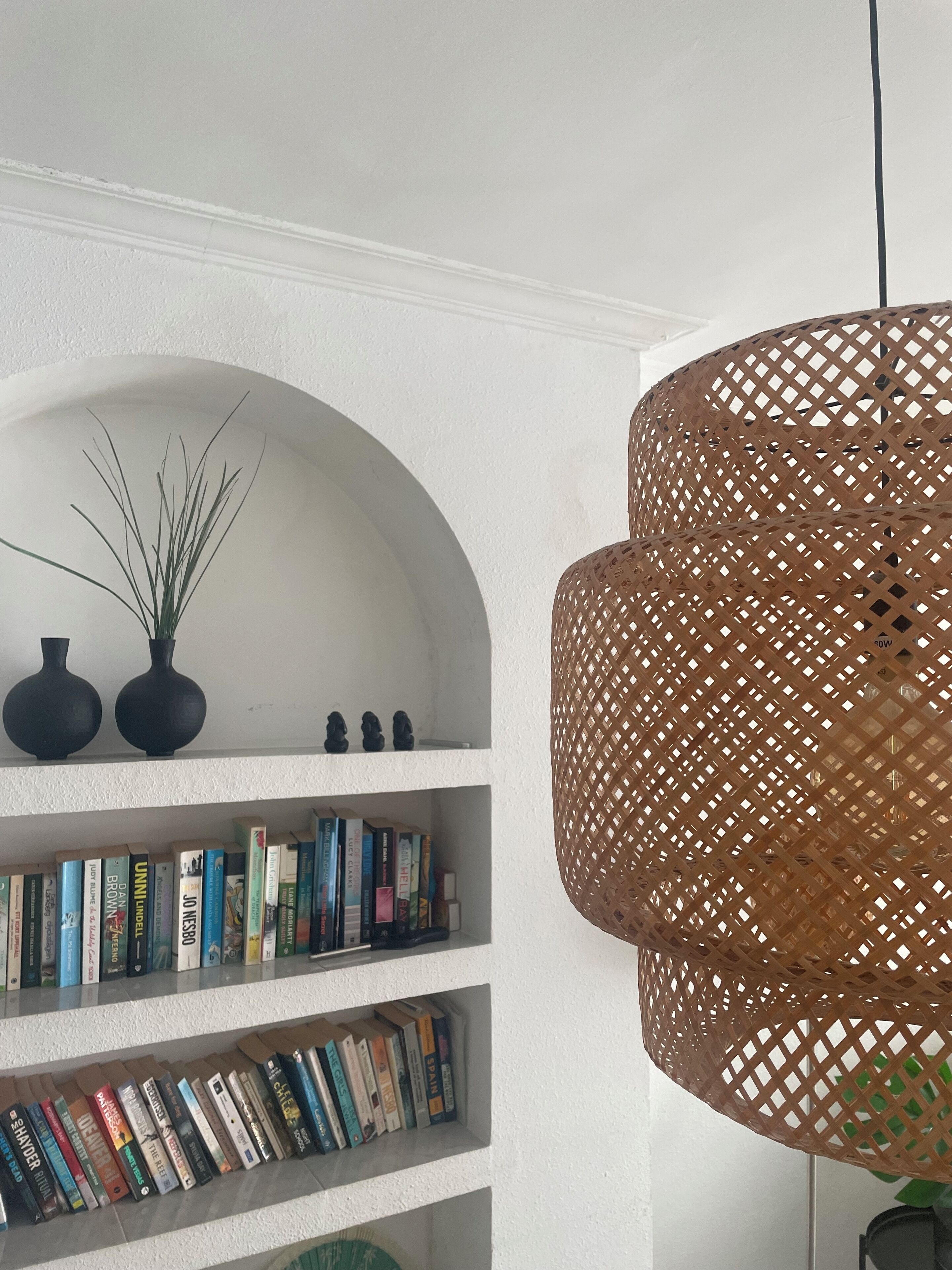 A bookshelf with books and decorative items, a woven pendant light, and a white wall with an arched niche.