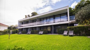 A modern two-story building with a flat roof, large windows, and a balcony. There are four lounge chairs in the front yard. - Studio north lower-level w/ kitchenett and patio (Santana)