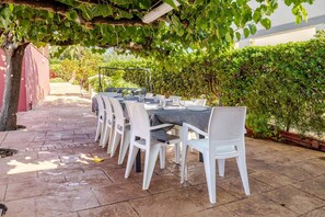 An outdoor dining area with white chairs and tables, covered by a pergola with green foliage.