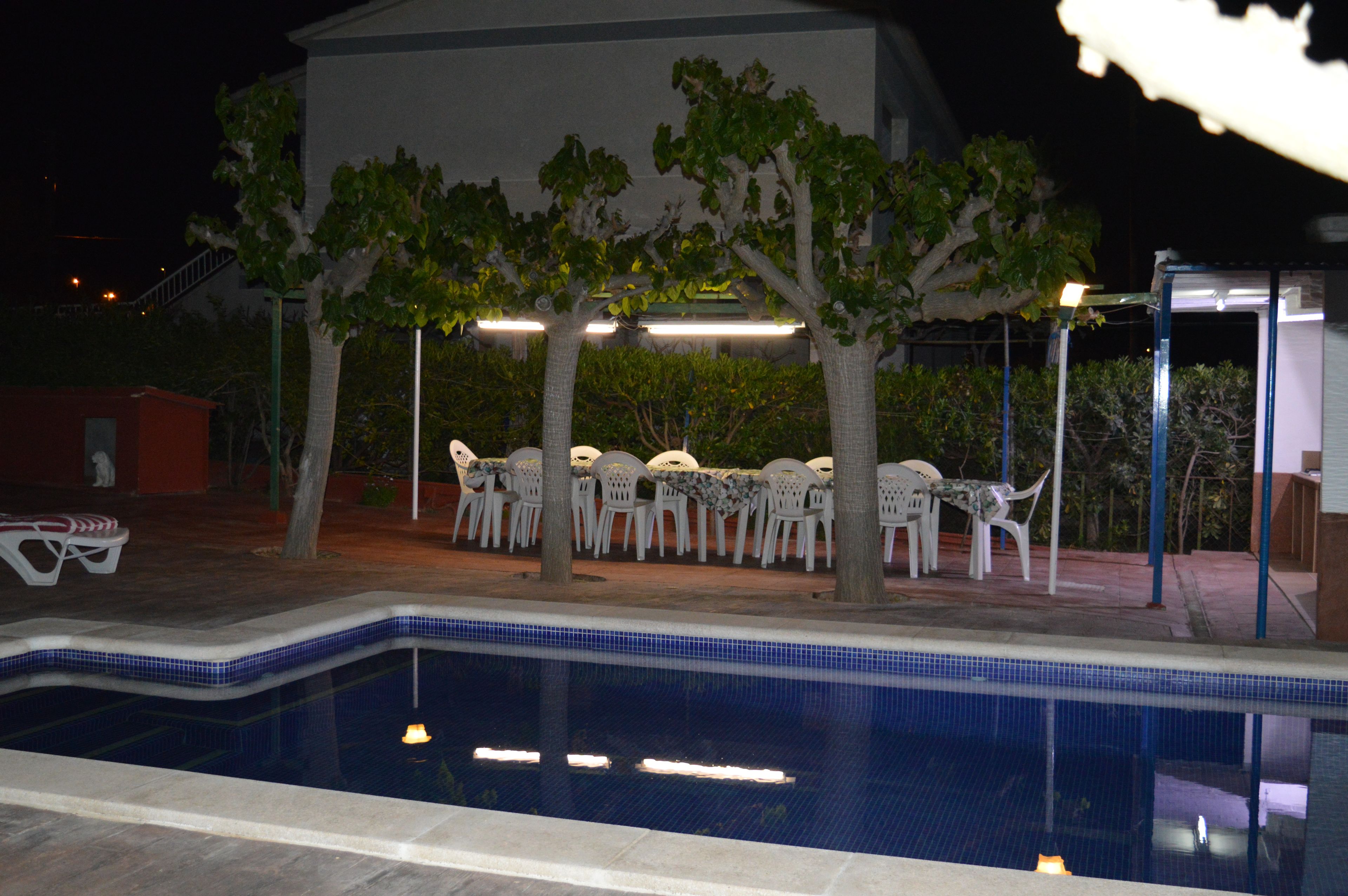 A swimming pool with a tiled edge, surrounded by white plastic chairs and tables under trees.