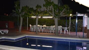 A swimming pool with a tiled edge, surrounded by white plastic chairs and tables under trees.
