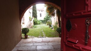 Ein Steinweg führt zu einer roten Tür, mit einem Ziegelbogen und Blick auf einen Garten sowie Gebäude im Hintergrund.
