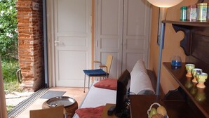 Ein gemütliches Zimmer mit einem Bett, einem Schreibtisch mit Computer, einem Sessel und einem kleinen Tisch mit Obstkorb. Eine Tür führt zu einem Balkon mit Blick auf Grünflächen.