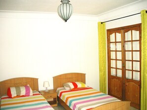 Quarto com duas camas, uma mesa de cabeceira com abajur, porta de madeira com painéis de vidro e luminária pendente.