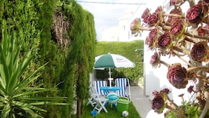 A small outdoor seating area with a table and chairs under a green and white umbrella, surrounded by lush greenery and a potted plant with purple flowers.