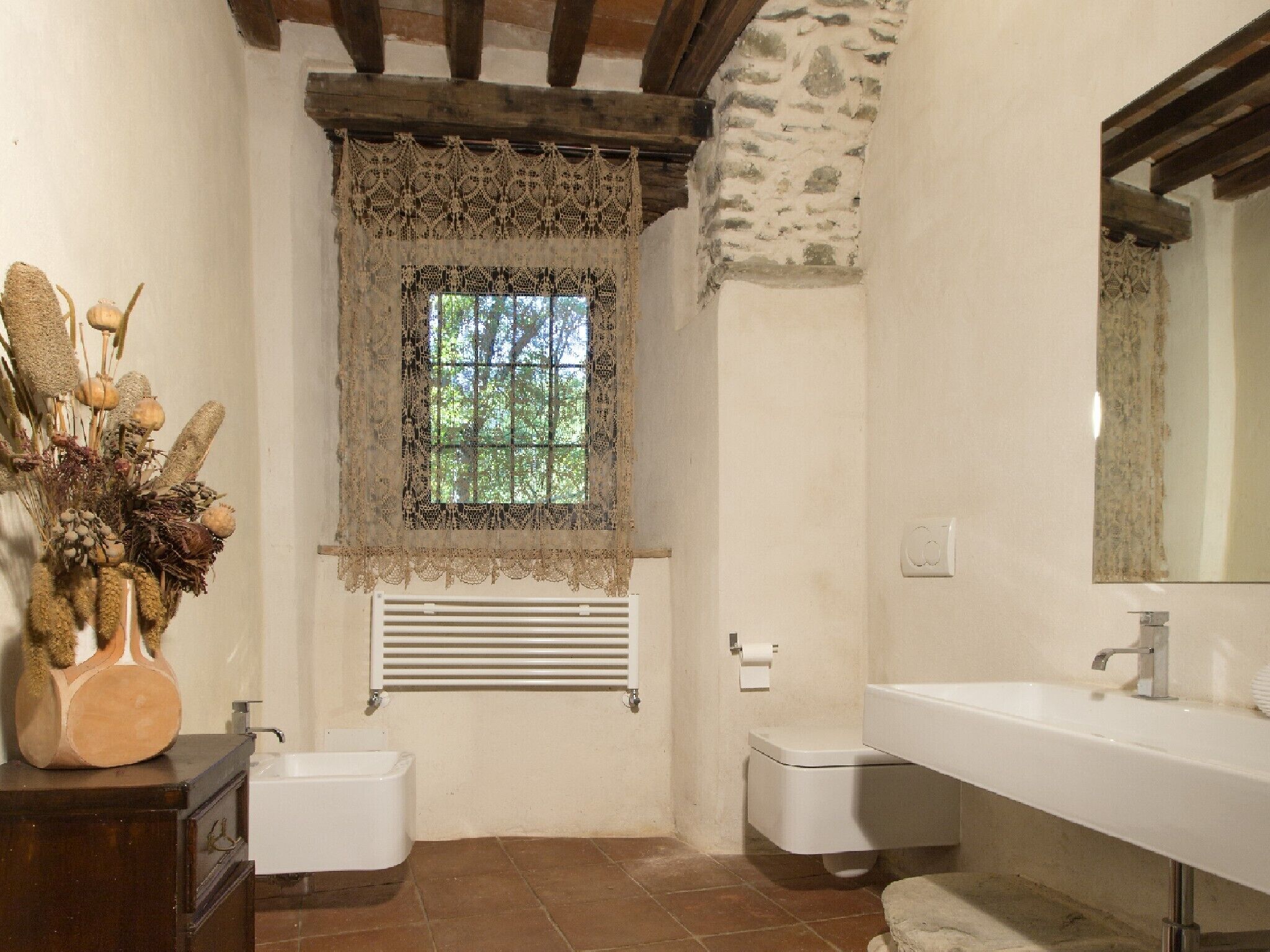 A bathroom with a rustic stone wall, a window with lace curtains, a white sink, and a wooden cabinet.