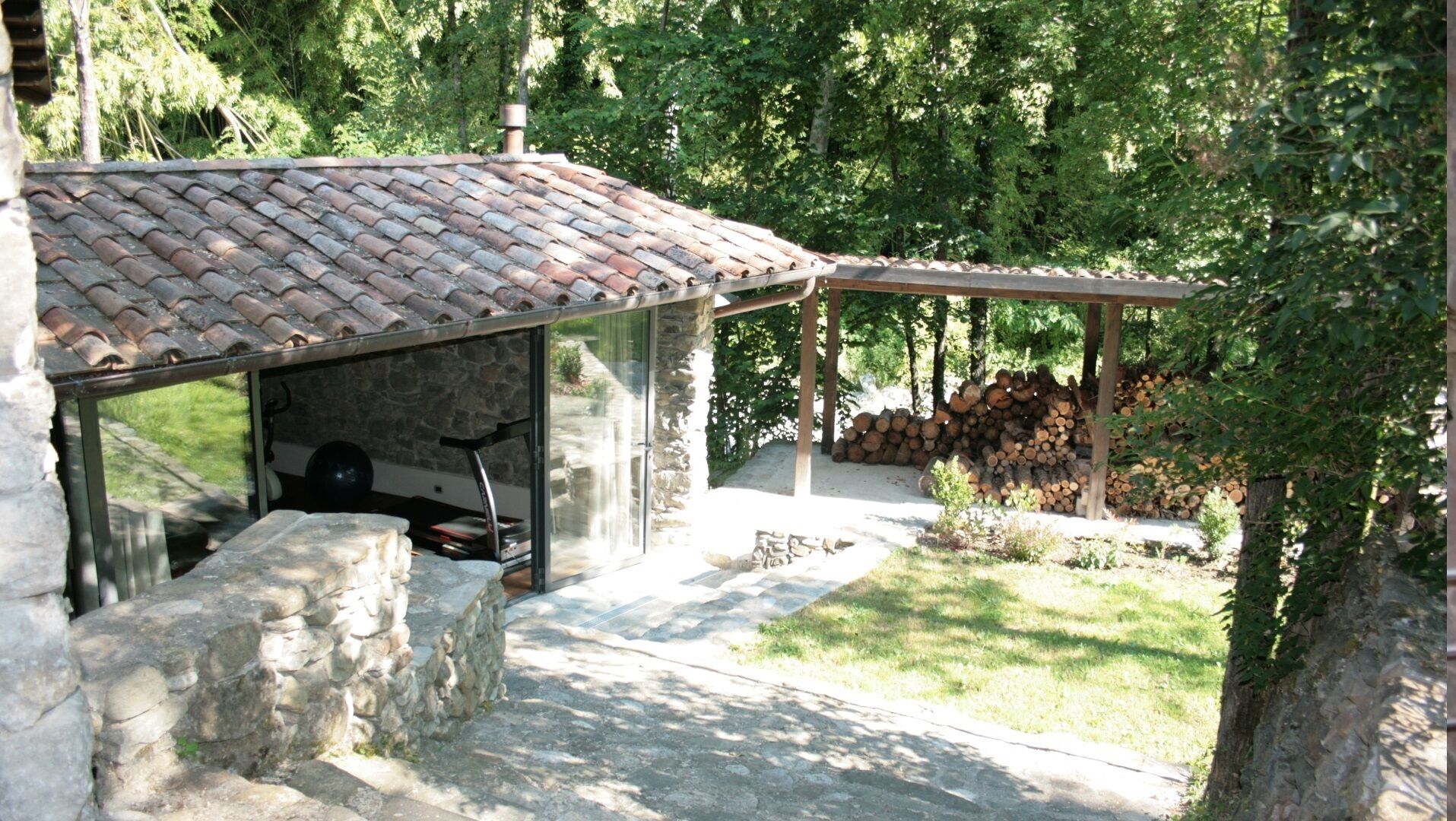 Un bâtiment en pierre avec un toit en tuiles, un espace extérieur couvert et une pile de bois de chauffage.