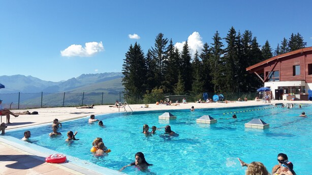 A swimming pool with people enjoying the water, surrounded by a building and trees, with mountains in the background.