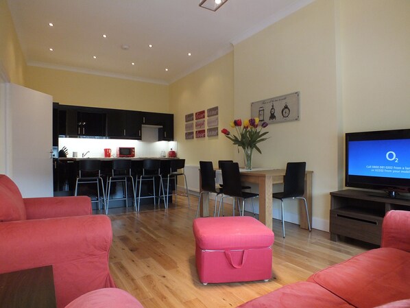 Una sala de estar moderna con piso de madera, un televisor, un otoman rosa y una cocina al fondo.