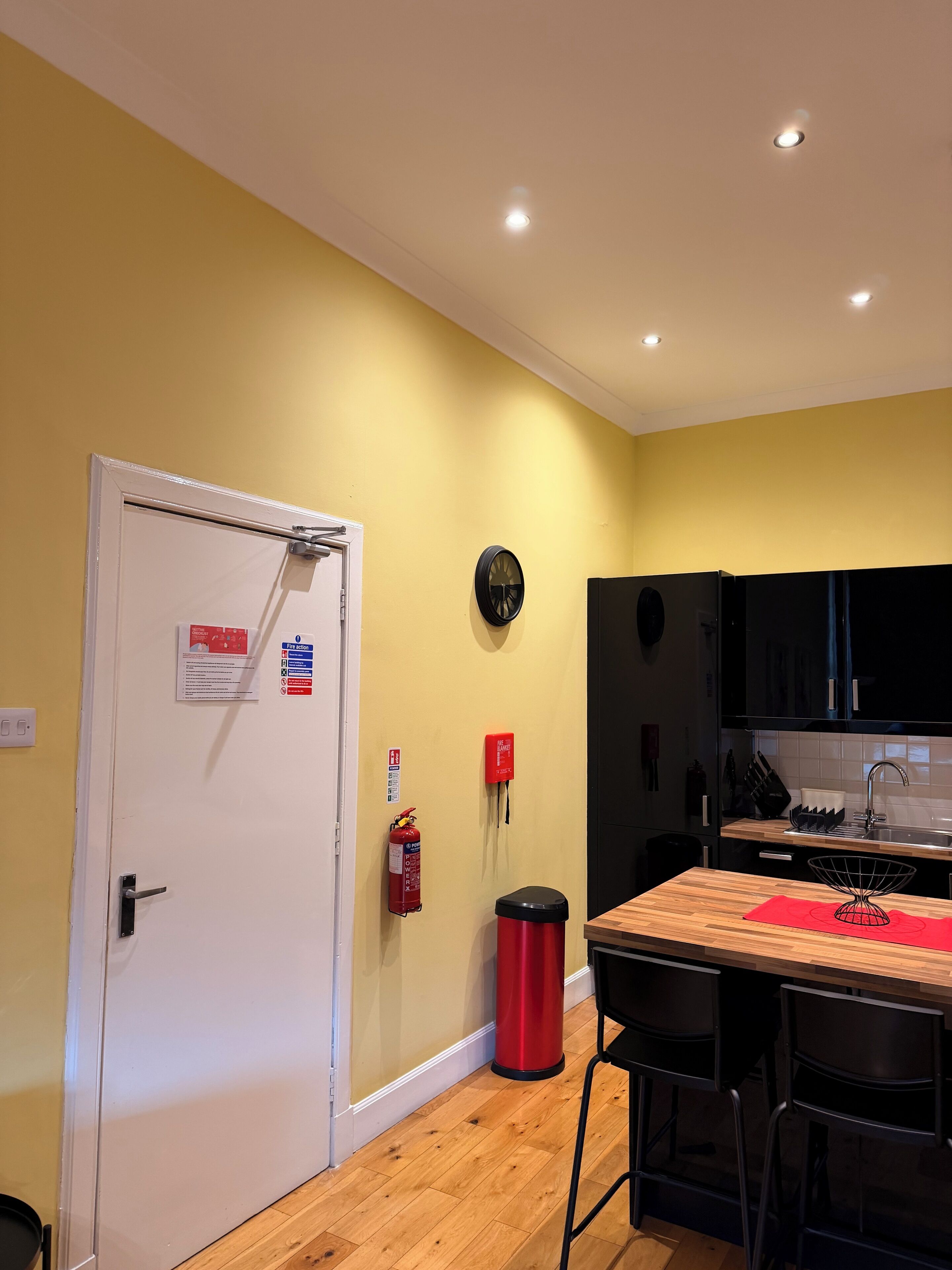 A kitchen with wooden flooring, a white door, a red fire extinguisher, and black cabinets.