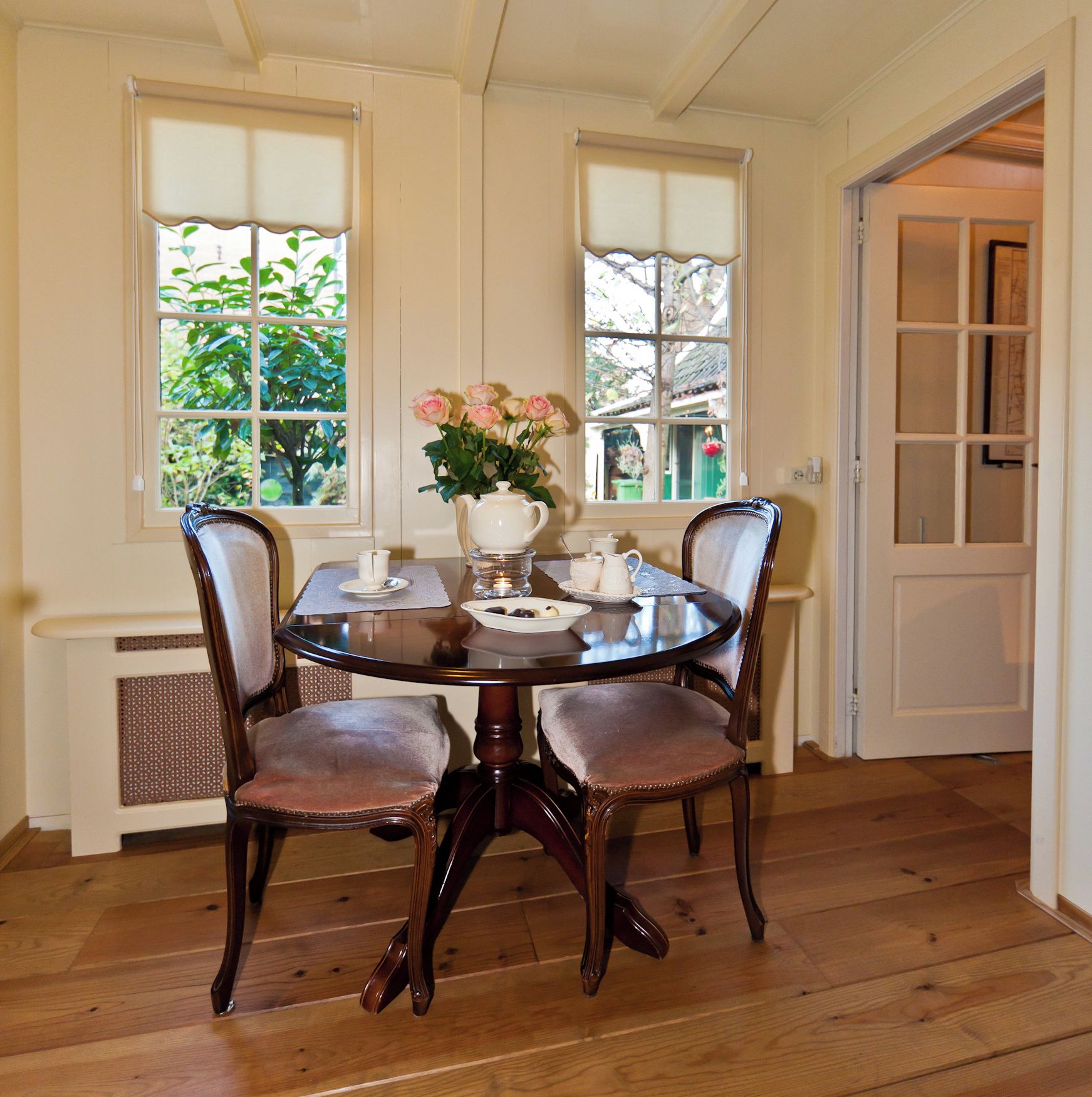 A dining room with a round table, four chairs, a vase with flowers, and a window with a view of greenery.