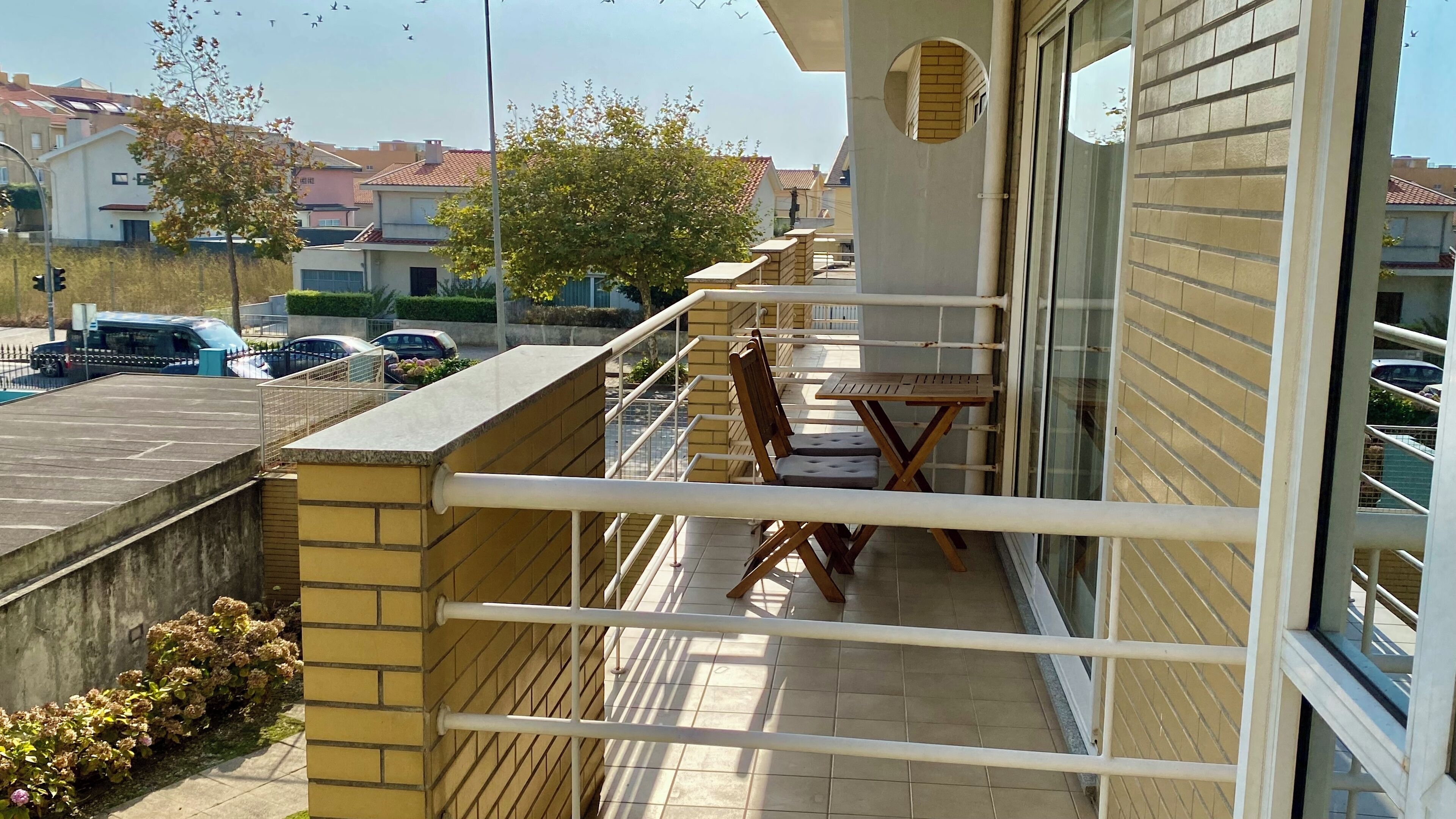 Uma varanda com mesa e cadeiras, vista para a rua e um edifício com telhado de cerâmica.