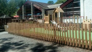 A building with a wooden fence, a playground, and a covered seating area. - Log Cabin On Shores Of Lake Windermere, Surrounded By Breathtaking Scenery (Windermere)