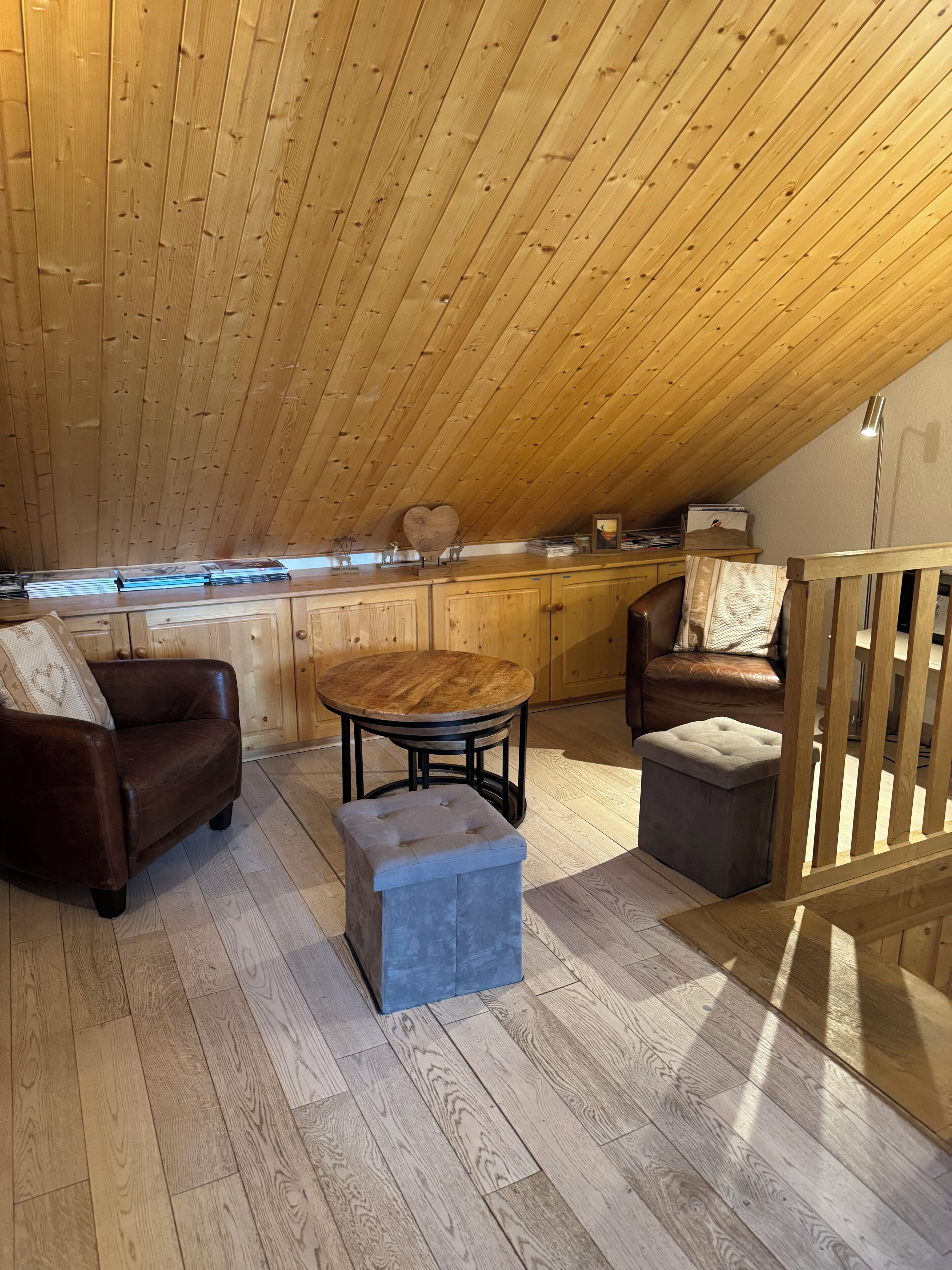 Ein gemütliches Schlafzimmer im Loft-Stil mit schrägem Dach, Holzboden, einem runden Holztisch, zwei Ledersofas und einem kleinen Hocker.