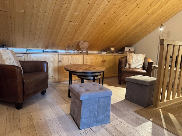 A cozy loft-style room with a wooden ceiling, a round dining table, a sofa, a bench, and a wooden railing.