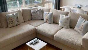 Una sala moderna con un sofá seccional color beige, una pequeña mesa de centro blanca y una mesa de comedor con sillas. Amplias ventanas ofrecen vistas al verde exterior.