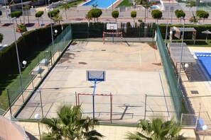 An outdoor basketball court with a net, surrounded by a fence, and palm trees in the foreground.